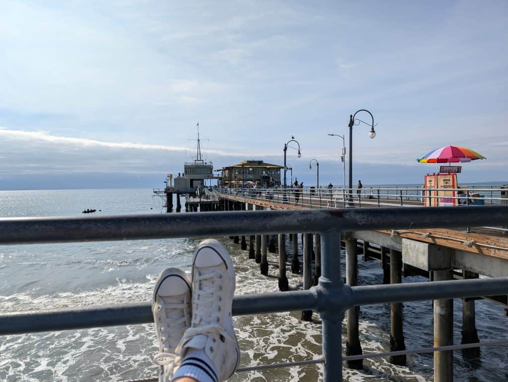 Blick auf den Santa Monica Pier mit dem Pazifik im Vordergrund und ausgestreckten Beinen auf der Promenade